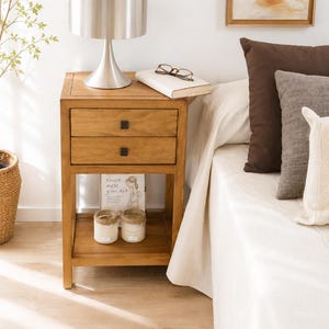 Peut inclure: Une table de chevet en bois brun clair avec deux tiroirs dotés de petits boutons carrés foncés. La table possède une étagère inférieure ouverte contenant deux petits bocaux en verre et un livre avec le texte Nueve meses y un día.