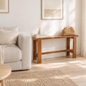 Könnte beinhalten: Eine rechteckige Holzbank mit vier stabilen Beinen und einer horizontalen Stützstrebe, die eine natürliche braune Holzmaserung aufweist. Die Bank steht vor einer weißen Wand und bietet eine Ablagefläche für Bücher und einen geflochtenen Korb.