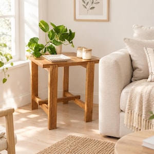 Op de afbeelding: Een vierkante houten bijzettafel met vier rechte poten en een onderste kruisverbinding. Het tafelblad bevat een wit vierkant decoratief dienblad en twee kleine glazen potjes met kurken deksels. De tafel is gemaakt van lichtbruin hout.