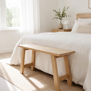 Pode incluir: Um banco de madeira de tom claro com quatro pernas anguladas e um assento retangular, posicionado ao pé de uma cama coberta com um edredão branco. O banco apresenta um acabamento de madeira natural lisa e um design de construção simples.