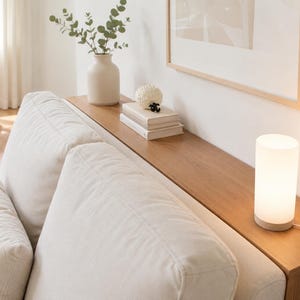 Puede incluir: Una lámpara de mesa cilíndrica con pantalla de vidrio esmerilado blanco y base de madera clara, que emite una luz cálida y suave. La lámpara está colocada sobre una consola de madera estrecha situada justo detrás de un sofá tapizado en color crema.