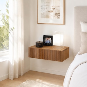 May include: A rectangular, wall-mounted floating bedside table crafted from light brown wood with a vertical slatted front design. The flat top surface holds a small black circular container, a black picture frame, and a square glass lamp.