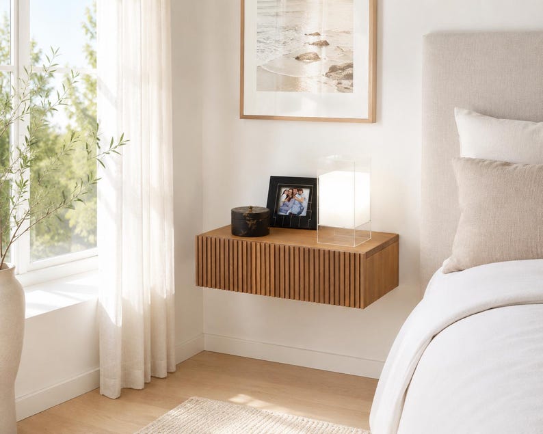 Puede incluir: Una mesita de noche flotante rectangular montada en la pared, hecha de madera de color marr&oacute;n claro con un dise&ntilde;o de listones verticales en el frente. La superficie superior plana sostiene un peque&ntilde;o recipiente circular negro, un marco de fotos negro y una l&aacute;mpara de cristal cuadrada.