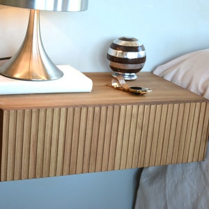 Puede incluir: Una mesita de noche flotante montada en la pared, hecha de madera de color marr&oacute;n claro con un panel frontal texturizado de listones verticales. La superficie rectangular sostiene una l&aacute;mpara de metal plateado, un libro blanco, un objeto esf&eacute;rico a rayas y un reloj dorado.