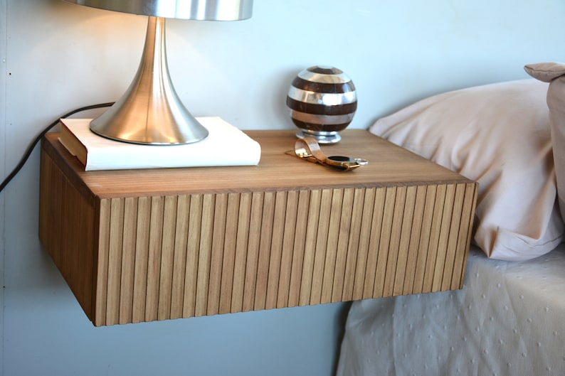 Puede incluir: Una mesita de noche flotante montada en la pared, hecha de madera de color marr&oacute;n claro con un panel frontal texturizado de listones verticales. La superficie rectangular sostiene una l&aacute;mpara de metal plateado, un libro blanco, un objeto esf&eacute;rico a rayas y un reloj dorado.