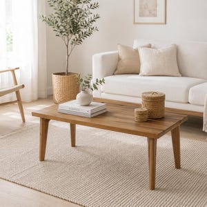 May include: A rectangular wooden coffee table with four tapered legs. On the table surface sits a stack of two white books, a small white ceramic vase with green leaves, and two round woven lidded baskets of different sizes in a natural tan color.