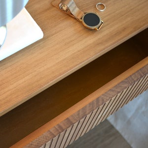 Puede incluir: Una mesita de noche flotante de madera marr&oacute;n claro con un caj&oacute;n abierto y un panel frontal de listones. Sobre la superficie superior hay un reloj de pulsera dorado con correa de malla y un anillo de metal plateado junto a una base de espejo blanca.