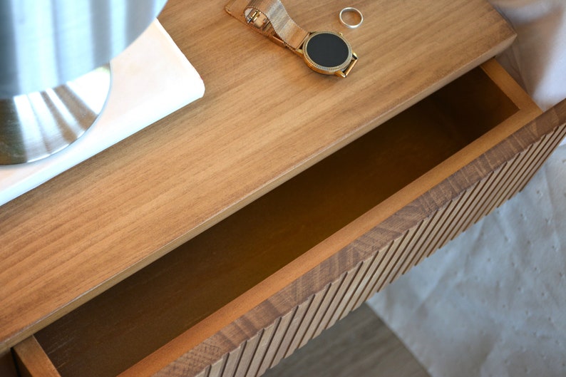 Puede incluir: Una mesita de noche flotante de madera marr&oacute;n claro con un caj&oacute;n abierto y un panel frontal de listones. Sobre la superficie superior hay un reloj de pulsera dorado con correa de malla y un anillo de metal plateado junto a una base de espejo blanca.