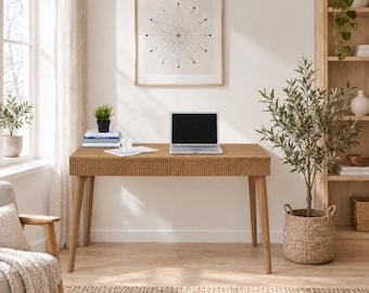 May include: A rectangular wooden desk with a fluted front drawer panel and four tapered legs. A silver laptop, a white mug, and a stack of books are placed on the surface. The desk is positioned in a room with a framed geometric print on the wall.