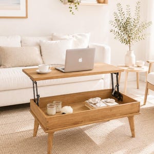 Peut inclure: Une table basse rectangulaire en bois dotée d'un mécanisme de plateau relevable. La table présente quatre pieds fuselés et un compartiment de rangement ouvert sous le plateau, contenant actuellement deux bocaux en verre et des livres.