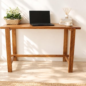 Könnte beinhalten: Ein rechteckiger Schreibtisch aus Holz mit naturfarbenem Finish, ausgestattet mit vier geraden Beinen und einer horizontalen Stützstrebe. Auf der Tischplatte stehen ein schwarzer Laptop, ein geflochtener Korb mit einer Grünpflanze und Bücher.