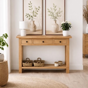 Puede incluir: Una consola de madera clara con tres cajones y un estante inferior, con dos cestas tejidas y tarros decorativos. Dos grabados botánicos enmarcados cuelgan sobre la mesa, y una lámpara blanca y una planta en maceta están encima. Una alfombra redonda de yute está en el suelo.