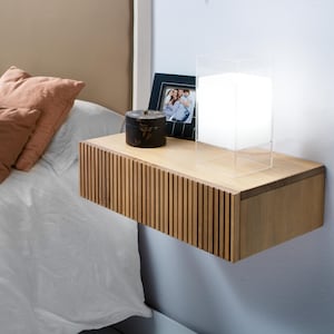 Może przedstawiać: Pływająca półka drewniana z szufladą, małym czarnym pudełkiem, oprawionym zdjęciem i przezroczystą lampą akrylową. Półka jest zamontowana na jasnoniebieskiej ścianie.