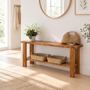 Pode incluir: Uma mesa consola retangular de madeira com acabamento castanho natural, com quatro pernas robustas e uma prateleira inferior. A mesa contém uma planta em vaso branco, uma mala de mão entrançada e dois cestos retangulares na prateleira.