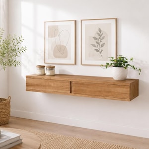 Könnte beinhalten: Ein rechteckiges, hellbraunes schwebendes Wandregal aus Holz mit zwei Schubladen und vertikalen Griffen. Das Regal ist an einer weißen Wand unter zwei gerahmten Kunstdrucken montiert, von denen einer abstrakte Formen und der andere ein Blatt zeigt.