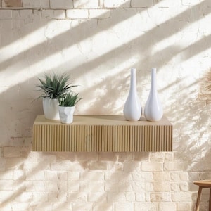 Może przedstawiać: Jasna drewniana półka z żebrowanym wzorem, zamontowana na białej ceglanej ścianie. Półka jest ozdobiona dwoma białymi ceramicznymi wazonami i dwoma doniczkami z roślinami.