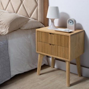 Op de afbeelding: Een nachtkastje van licht hout met twee laden en een lattenfront. Het nachtkastje heeft vier poten en staat naast een bed met een wit hoofdeinde.