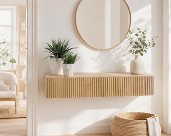 Puede incluir: Una consola flotante de madera clara montada en la pared con un diseño de listones verticales en el frente. Sobre la consola hay un gran espejo redondo con un marco de madera fino. Dos plantas verdes en macetas y un jarrón beige con eucalipto.