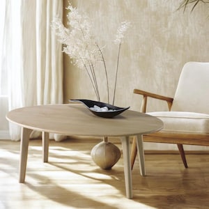 Op de afbeelding: Een ovale salontafel van licht hout met een zwarte, blaadervormige kom erop. De kom is gevuld met witte stenen. De tafel heeft vier poten en staat op een lichte houten vloer. Een witte stoel met een licht houten frame staat op de achtergrond.