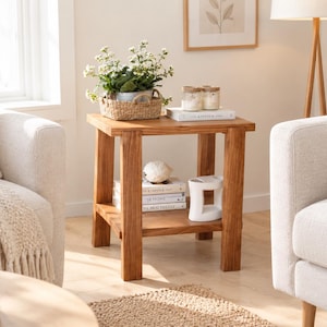 Peut inclure: Une table d'appoint carrée en bois avec quatre pieds robustes et une étagère inférieure. Le plateau supporte un panier tressé avec des fleurs blanches et deux bougies sur un livre. L'étagère inférieure contient des livres, un objet blanc et un vase.