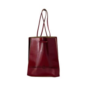 May include: A deep red leather tote bag with a smooth finish, featuring two thin shoulder straps and a drawstring closure at the top. The lower section of the bag displays a horizontal band of intricate, laser-cut leaf and floral patterns.