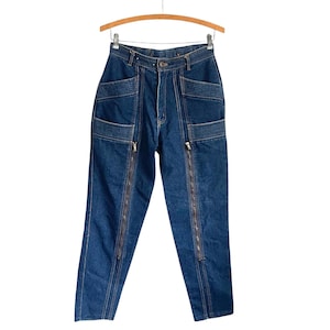 May include: Dark blue denim jeans with a high waist and multiple pockets. The jeans feature two vertical zippers running down the front of each leg. The jeans are hanging on a wooden hanger.