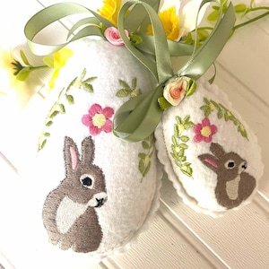 May include: Two white felt Easter eggs with embroidered bunnies and floral designs. The eggs are tied together with a green ribbon and have a small pink flower embellishment.