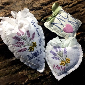 Könnte beinhalten: Drei weiße herzförmige Säckchen mit Lavendelstickerei und dem Wort "Mama" auf jedem. Die Säckchen haben verschiedene Designs, darunter ein Lavendelstrauß, ein Herz und die Worte "Wir lieben dich".
