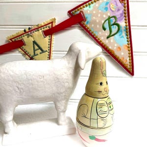 May include: A white ceramic sheep figurine stands next to a painted wooden bunny toy. A banner with the letters "A" and "B" is strung across the top. The banner is made of fabric with floral patterns and red trim.
