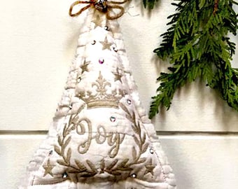 May include: A white fabric Christmas tree ornament with a brown twig trunk. The ornament is decorated with gold embroidery, including a crown, stars, and the word "Joy".