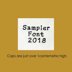 Puede incluir: Una etiqueta de tela blanca con texto negro que dice "Sampler Font 2018". El texto está cosido en la tela. La leyenda debajo de la etiqueta dice "Las mayúsculas tienen poco más de 1 centímetro de alto."