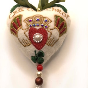 Puede incluir: Adorno de corazón de fieltro blanco con un diseño Claddagh bordado, un trébol verde y el texto "Gra o Chroi". El adorno está decorado con cuentas rojas, doradas y blancas y un colgante de cristal.