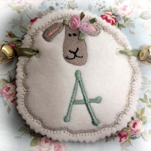 May include: A white felt sheep with a green letter A embroidered on it. The sheep has a brown face with pink flowers on its head. The sheep is attached to a bell on each side.