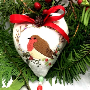Op de afbeelding: Een hartvormig, wit stoffen ornament met een geborduurd roodborstje-ontwerp, rode strik en kleine gouden bel. De vogel heeft een bruine rug, een rode borst en is omgeven door een groene krans met rode bessen. Het ornament is versierd met kralen en omgeven door dennentakken.