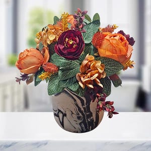 May include: A floral arrangement featuring deep burgundy and vibrant orange artificial flowers, including peonies and hydrangeas, accented with green leaves and small textured sprigs, all contained within a rounded ceramic vase with a black and white marble pattern.