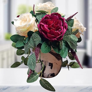 May include: A floral arrangement featuring a large deep burgundy peony, several cream-colored roses, and dark green leaves with visible veins. The flowers are contained in a rounded vase with a marbled black and white pattern and accented by thin purple sprigs.