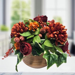 May include: A floral arrangement featuring deep red and burnt orange chrysanthemums, dark red roses, and small textured berries. The flowers are accented with green ivy leaves and are contained within a round, horizontally ribbed tan ceramic bowl.