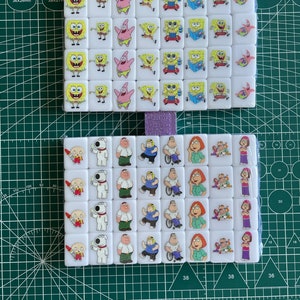 May include: A set of 38 white ceramic dominoes featuring colourful cartoon images of characters from the animated television series SpongeBob SquarePants and Family Guy. The dominoes are arranged in a grid pattern on a green cutting mat.