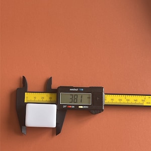 May include: A white rectangular object is measured with a black digital caliper. The caliper displays a measurement of 38.1 mm. The background is a solid orange-brown color.
