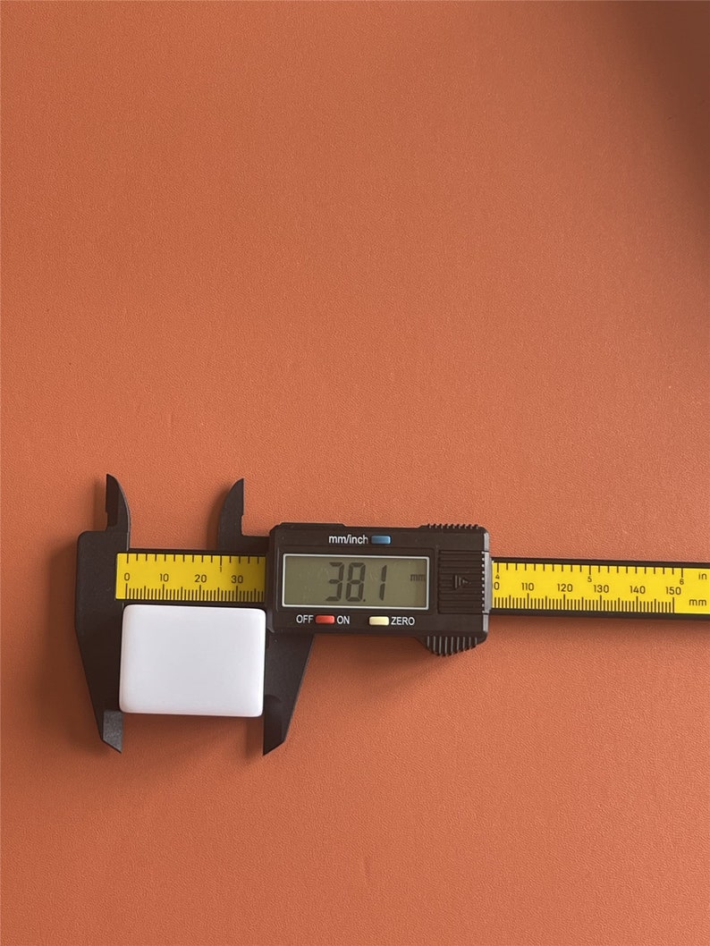 May include: A white rectangular object is measured with a black digital caliper. The caliper displays a measurement of 38.1 mm. The background is a solid orange-brown color.