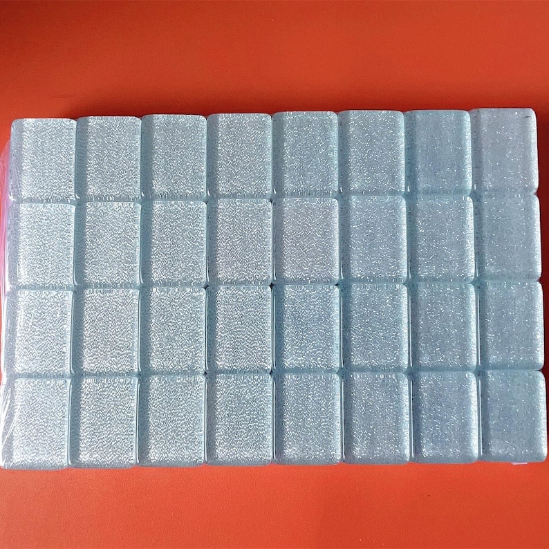 May include: A package of small, rectangular, light blue glass tiles. The tiles have a textured surface and are arranged in a grid pattern. The background is a solid orange color.