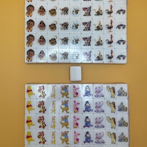 May include: Two sets of white Mahjong tiles featuring colorful cartoon character designs. The top set includes characters from the movie Moana, while the bottom set features Winnie the Pooh and friends. A single spare tile is centered.