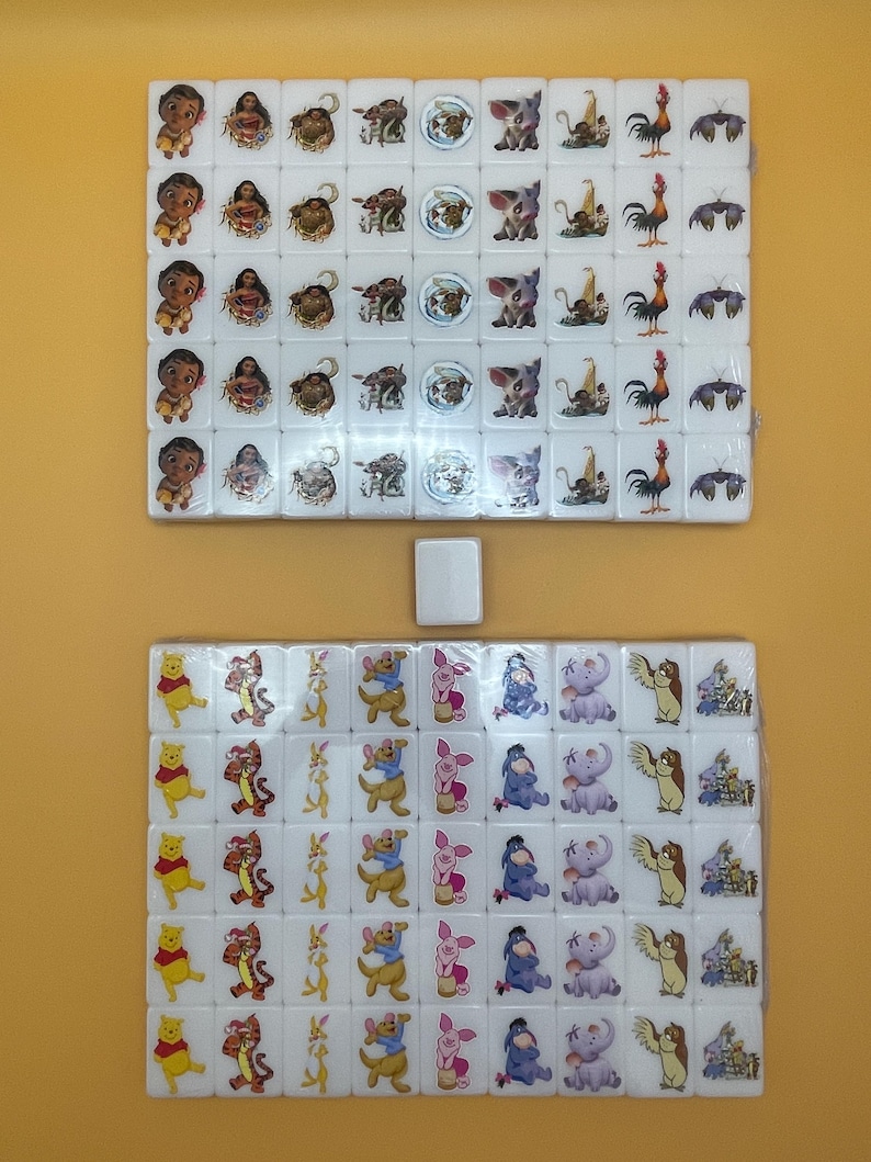 May include: Two sets of white Mahjong tiles featuring colorful cartoon character designs. The top set includes characters from the movie Moana, while the bottom set features Winnie the Pooh and friends. A single spare tile is centered.