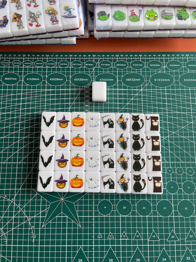 May include: A set of 16 white dominoes featuring Halloween-themed images. The images include a bat, a witch's hat, a pumpkin, a ghost, a grim reaper, a black cat, and a spooky tree. The dominoes are arranged in a grid pattern on a green cutting mat.