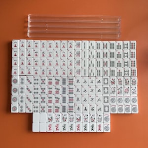 May include: A set of white mahjong tiles with red and black markings. The tiles are arranged in a grid pattern and include numbers, symbols, and characters. The tiles are on a brown surface.