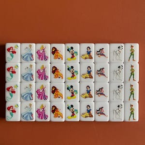 May include: A set of 36 white dominoes featuring colourful Disney character illustrations. The characters include Ariel, Cinderella, Snow White, Mickey Mouse, Simba, and Peter Pan.