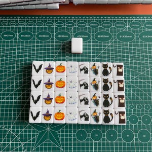 May include: A set of 36 white dominoes with Halloween-themed images. The dominoes feature images of bats, witches, pumpkins, ghosts, and skeletons. The dominoes are arranged in a grid pattern on a green cutting mat.