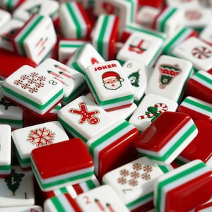 Op de afbeelding: Een verzameling rechthoekige Mahjong-tegels met kerstthema-illustraties, waaronder een peperkoekmannetje, een kerstman met de tekst JOKER, een sneeuwvlok, zuurstokken, pepermuntjes, een bel en een kerstboom in rood, groen en wit.