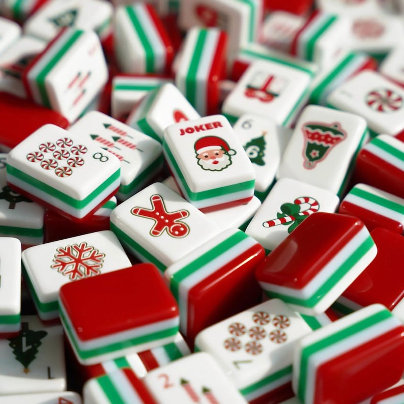Op de afbeelding: Een verzameling rechthoekige Mahjong-tegels met kerstthema-illustraties, waaronder een peperkoekmannetje, een kerstman met de tekst JOKER, een sneeuwvlok, zuurstokken, pepermuntjes, een bel en een kerstboom in rood, groen en wit.