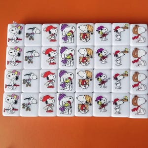 May include: A set of 36 white dominoes with colorful images of Snoopy and Woodstock. Each domino features a different image of the characters, including Snoopy wearing a hat, holding a bird, and wearing a scarf.